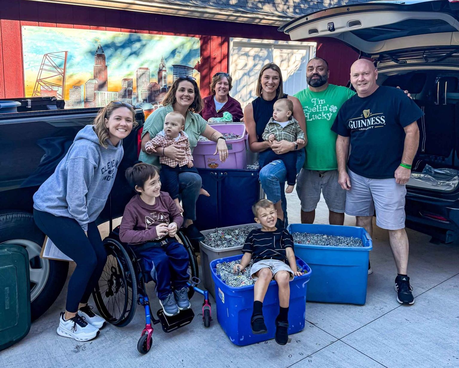 group of people including four children, in front of an open car hatch. They are donating four large plastic bins ofpull tabs