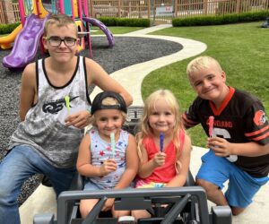 Two girls in power wheels car, boys on either side, on a playground