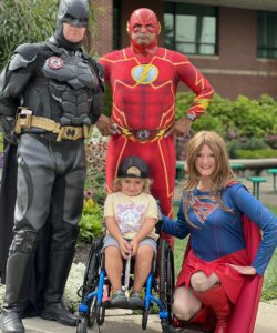 Costumed characters of Batman, Supergirl and one other with child in wheelchair