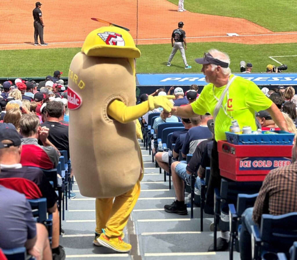 Joe shakes hands with a person in a hotdog outfit. they are on the stadium stairs.