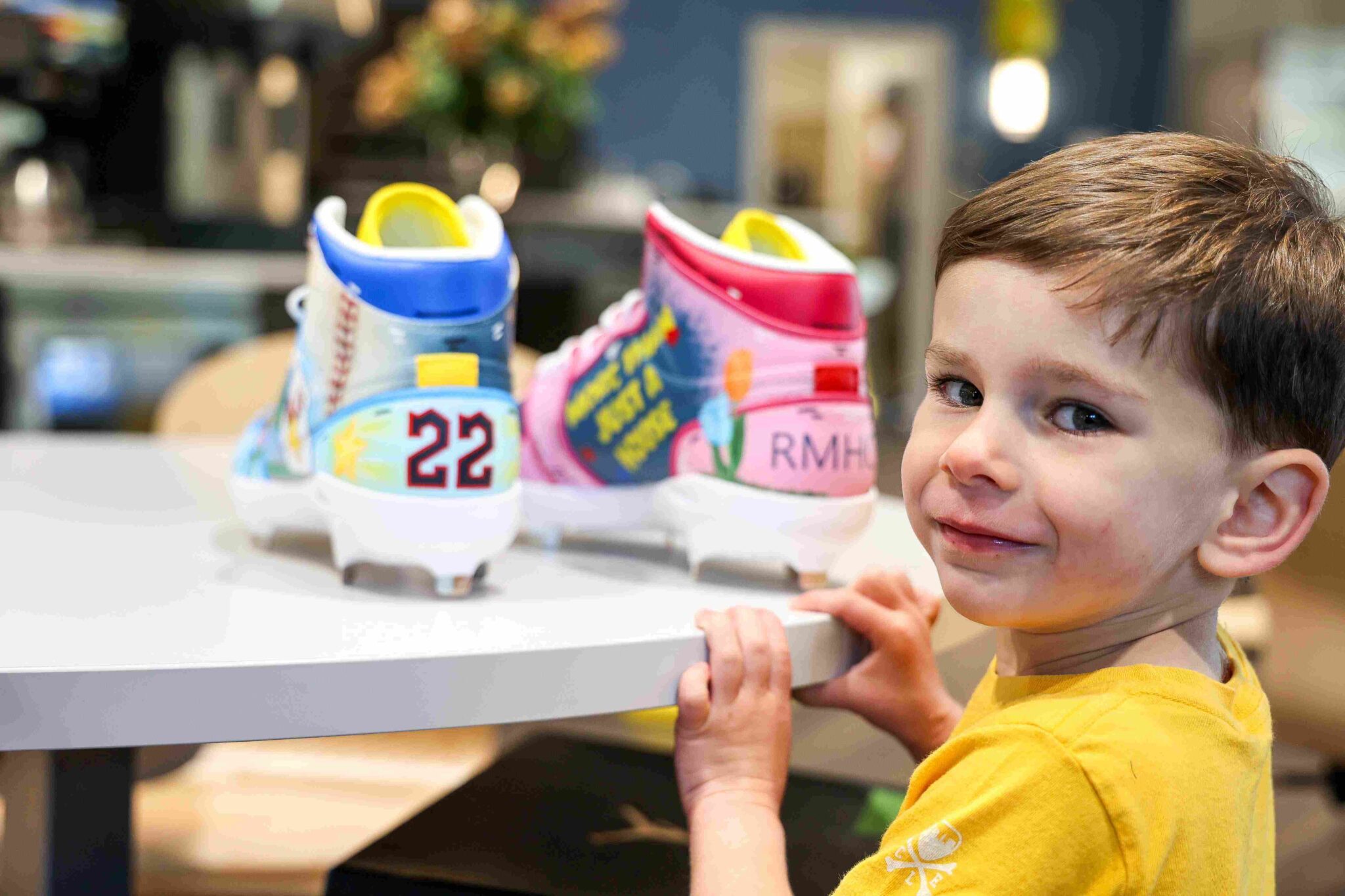 Small child stands in front of table. Hand-painted baseball cleats rest on table.