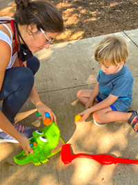 RMHC client kneeling on sidewalk with child while they play with toys.